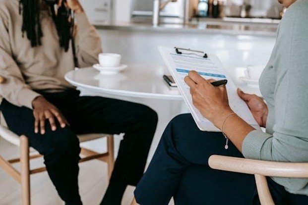 Two people sitting across from each other at a table while one holds a clipboard and a pen